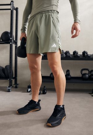 Shorts de sport verts avec une ceinture et des fentes latérales, associés à des chaussures noires avec une semelle gum, dans un environnement de salle de sport.
