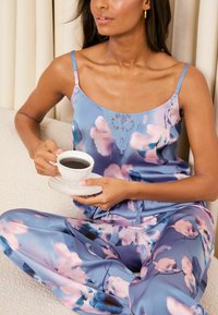 Set de pijamale din satin, albastru, cu modele florale roz. Bretele spaghetti și accent din dantelă. Model ținând o ceașcă de cafea pe o canapea bej.