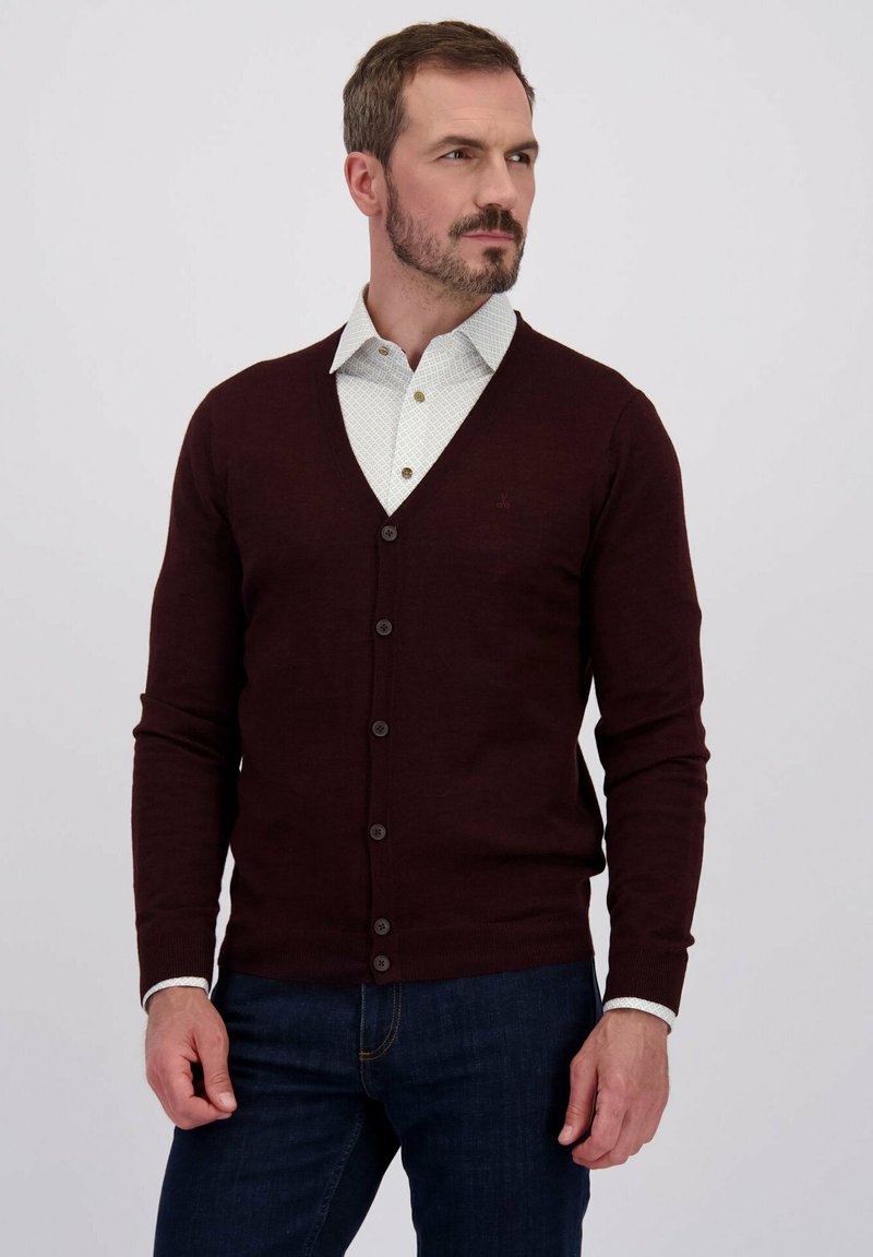 Man met een baard in een donker bordeauxrode gebuttonde cardigan over een wit met patroon shirt en donkerblauwe spijkerbroek, kijkend naar rechts.