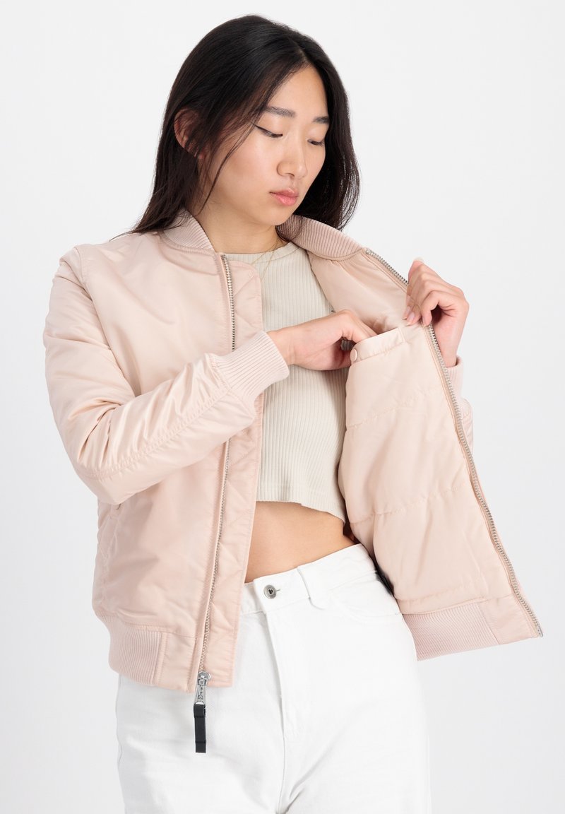 Helle rosa Bomberjacke aus glattem Stoff, mit Reißverschluss vorne, gerippten Bündchen und einer Innentasche. Über einem hellen Oberteil getragen.