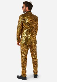 Dzeltena un melna raksta uzvalks ar zvaigžņu dizainu un spīdīgu, metālisku apdari, apvienots ar melniem uzvalka apaviem.