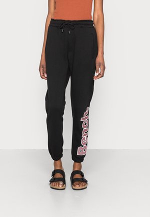 Personne portant un pantalon de jogging noir avec des poignets élastiques et un grand logo « Bench » blanc et rouge sur la jambe gauche, associé à des sandales noires et un haut orange.