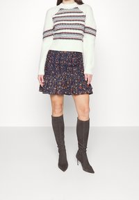 Femme portant un pull blanc à motifs, une jupe marine à volants fleuris et des bottes grises en daim montant jusqu'aux genoux avec talons, debout devant un fond blanc.