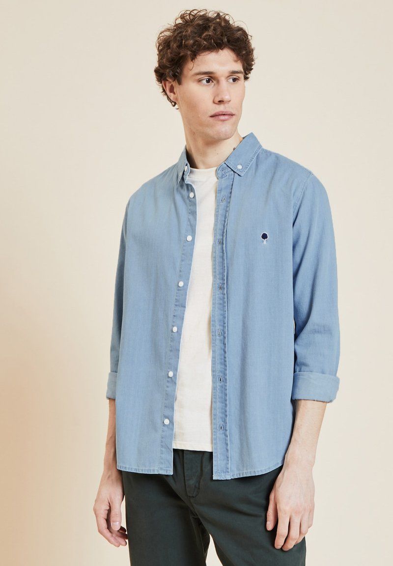 Camicia in denim blu chiaro con bottoni, collo arrotondato, maniche lunghe arrotolate fino ai gomiti e piccolo logo di un albero sul petto.