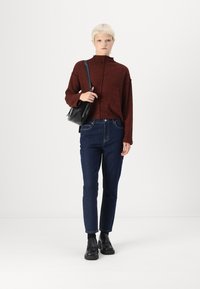 Sarkans augstais kakls džemperis, augsto vidukli tumšās džinsās un melni potīšu zābaki. Modelis tur nelielu melnu somu. Minimālisma fons.