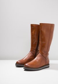 Bottes en cuir marron montant jusqu'au genou, avec une finition lisse, un bout arrondi, une fermeture éclair sur le côté et une semelle en caoutchouc plate. Accents texturés près de la cheville.