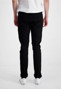 Personne portant un pantalon noir à revers et des baskets blanches, debout de dos face à l'appareil photo sur un fond léger uni.