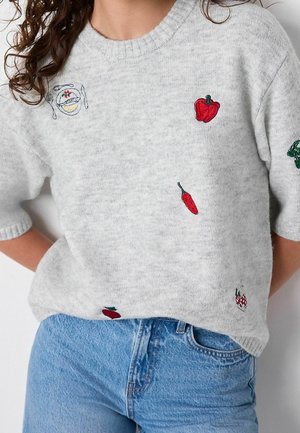Suéter corto de color gris claro con pimientos rojos bordados, un plato con cubiertos y pequeñas verduras verdes, combinado con jeans azules.