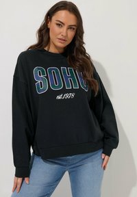 Sudadera negra con la inscripción "SOHO" en gran formato y detalle de cuadros. Con mangas largas y corte relajado, combinada con jeans de color azul claro.