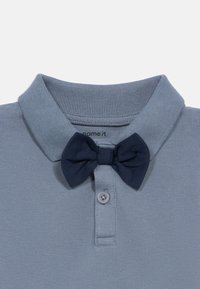 Μπλε πλενόμενο πουκάμισο πόλο με σκούρο μπλε υφασμάτινη γραβάτα κασκόλ προσαρτημένη κάτω από τον γιακά και ένα μόνο κουμπί στο πλύντ.