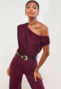Burgunderfarbener Jumpsuit mit schulterfreiem Design, drapiertem Stoff, gerippter Ärmeldetail und einem schwarzen, strukturierten Gürtel mit einer goldfarbenen Schnalle.