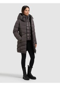 Abrigo tipo puff en gris oscuro con capucha, con cierre de cremallera en la parte delantera y bolsillos laterales, diseño acolchado y corte a media longitud; combinado con leggings negros y botas.
