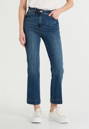 Jean en denim bleu foncé taille haute avec une jambe droite, un délavage subtil et des ourlets retroussés, associés à des baskets blanches.