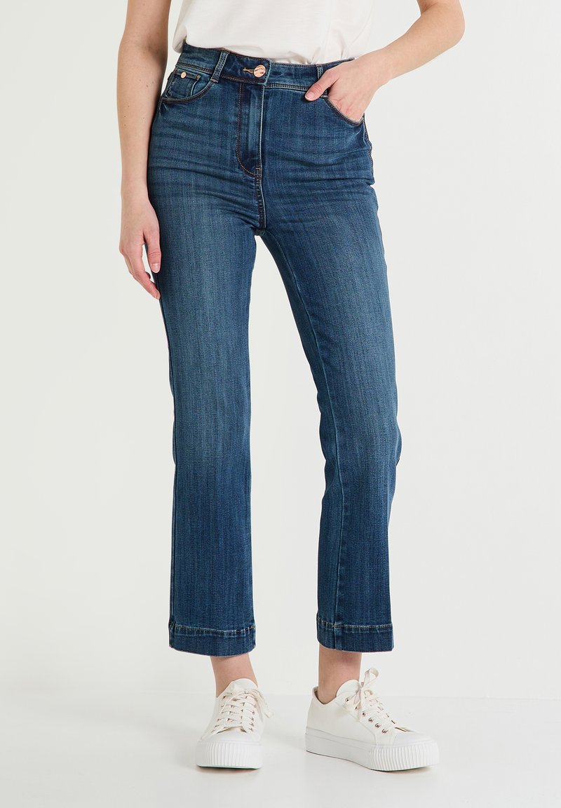 Jeans de mezclilla azul oscuro de cintura alta con pierna recta, ligeros desgastes y puños enrollados, combinados con zapatillas blancas.