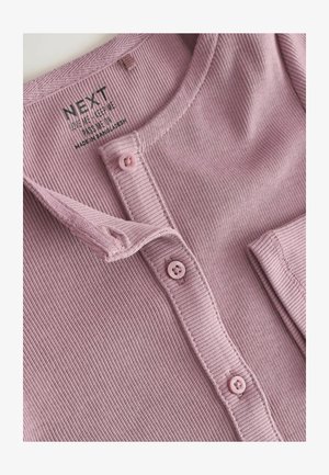 Lichtroze geribbelde top met een knoopsluiting aan de voorkant, voorzien van twee roze knopen en een zachte, textuurvolle stof. Inclusief een label met verzorgingsinstructies.