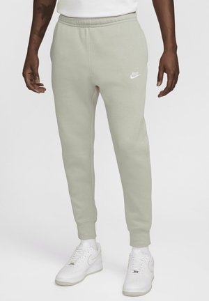 Person iført lysegrå Nike træningsbukser med elastiske manchetter og hvide Nike sneakers på en ensfarvet hvid baggrund.