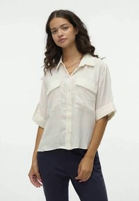 Blusa leve de creme com mangas curtas dobradas, fecho frontal com botões e dois bolsos no peito; apresenta uma textura subtil e um corte descontraído.