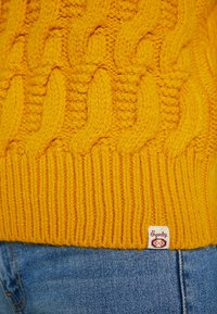 Gul stickad tröja med kabelmönster och ribbat nederkant; inkluderar en liten logotypetikett i det nedre högra hörnet.