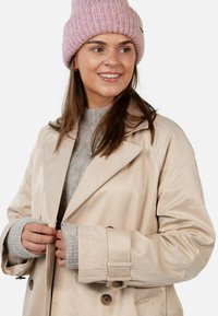 Young woman smiling, wearing a pink knit beanie, gray sweater, and beige trench coat, adjusting the coat buttons.