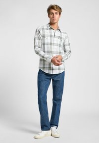 Camicia a quadri bianca, verde e blu navy, con colletto button-down e due tasche sul petto, abbinata a jeans blu e sneakers bianche.