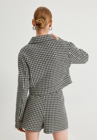 Zwart-wit houndstooth-gepatenteerde jas met lange mouwen en een verkorte zoom, gecombineerd met bijpassende shorts, die een gevarieerd stof tonen.