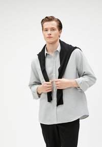 Lichtgrijs button-up shirt met een puntige kraag, gecombineerd met een zwarte trui die over de schouders is gedrapeerd. Zwarte broek maakt de outfit compleet.
