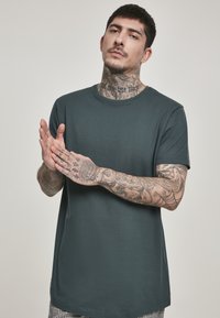Tamnozeleni pamučni majica s okruglim izrezom, kratkim rukavima i ravnim donjim rubom. Model ima vidljive tetovaže na rukama.