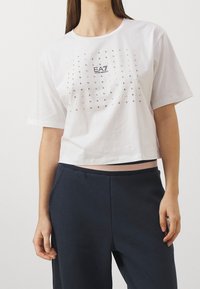 Bílý krátký bavlněný tričko se strukturovaným designem, zdobené kovovými nýty v mřížkovém vzoru a logem na přední straně.