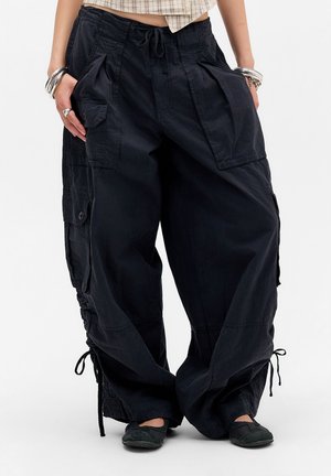 Femme portant un pantalon cargo noir ample avec de grandes poches et des cordons aux chevilles, associé à des chaussures noires et des bracelets argentés.