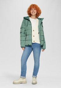 Gevulde groene jas met een capuchon, ritsen en elastische mouwen. Gemaakt boven een crèmekleurige trui en een blauwe jeans, gecombineerd met lichtbeige enkellaarsjes.