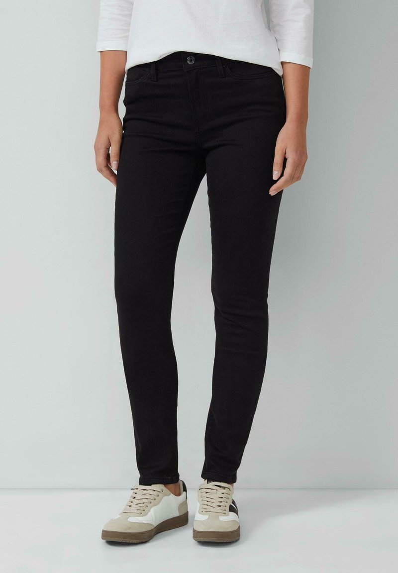 Jean slim noir en denim au toucher lisse, avec poches avant et fermeture à bouton. Porté avec des baskets blanches et beiges.