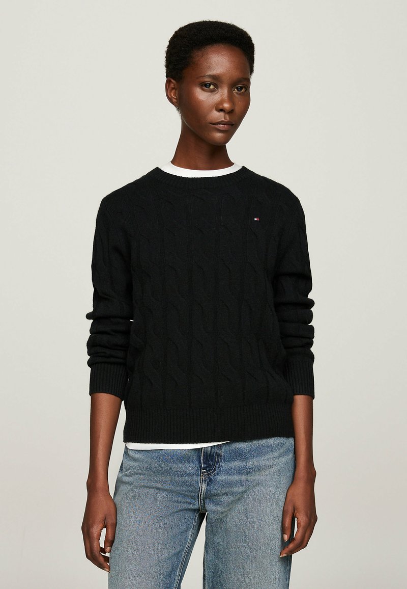 Pull-over en tricot noir avec un col rond et des poignets côtelés, porté sur une chemise blanche, complété par un jean bleu clair.