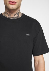 Camiseta negra de algodón de manga corta con cuello redondo y un pequeño logo blanco de "VANS" en el pecho. Textura suave, ajuste regular.