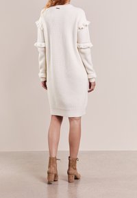 Biała sweterkowa sukienka z prążkowanej dzianiny, z falbaniastymi akcentami na rękawach i u dołu. Połączona z beżowymi botkami za kostkę na słupku z detalem w postaci kokardy.