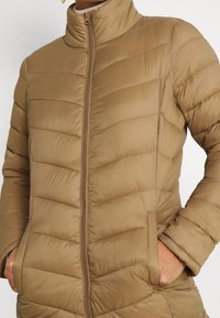 Tan, isolerad pufferjacka med hög krage, dragkedja och sidofickor; har horisontell quiltad sömnad och en mjuk textur.