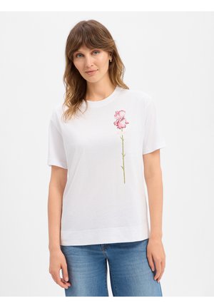 Femme aux cheveux châtain clair portant un T-shirt blanc avec un motif vertical de fleur rose sur la poitrine gauche et un jean bleu.