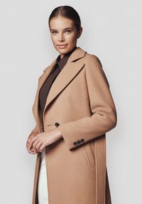 Beżowy wełniany płaszcz o strukturalnym kroju, z klapami i bocznymi kieszeniami. Posiada czarne guziki na rękawach oraz gładką, miękką teksturę.