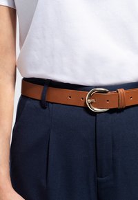Ceinture en cuir marron avec une boucle circulaire dorée, portée sur un pantalon marine, associée à un t-shirt blanc uni. Tissu texturé avec des plis.