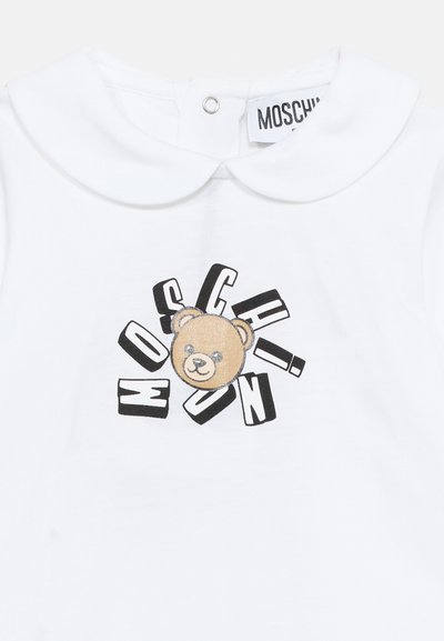 Λευκό παιδικό πουκάμισο με στρογγυλή γιακά που έχει το πρόσωπο ενός καφέ λούτρινου αρκούδου, περιτριγυρισμένο από διάσπαρτα μαύρα και λευκά γράμματα που σχηματίζουν το "MOSCHINO".