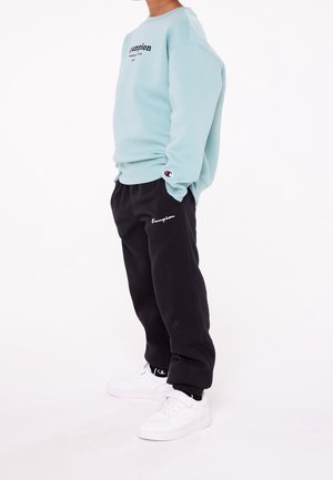 Lichtblauwe sweatshirt met zwart "Champion" logo, gecombineerd met zwarte trainingsbroek met wit "Champion" logo en witte sneakers.