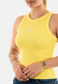 Top de tirantes amarillo con cuello redondo, que presenta una pequeña etiqueta de logo. La modelo lleva vaqueros de mezclilla y se complementa con pulseras y un reloj.