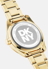 Orologio in acciaio inox dorato con cinturino metallico, caratterizzato da un quadrante cronografo grigio tondo e un logo DKNY ben visibile sul retro. Resistente all'acqua fino a 5 ATM.