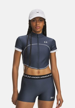 Vrouw met een witte pet, navyblauwe cropped zip-up top en bijpassende Under Armour short, staand tegen een effen achtergrond.
