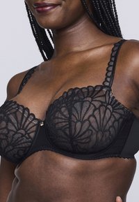 Reggiseno in pizzo nero con dettagli a motivo floreale, supporto con ferretto, bordi smerlati e spalline regolabili. Include un piccolo accento dorato.