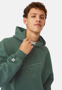 Grön hoodie med en mjuk textur, med en framsidestämpel "Ärlighet Regler" i upphöjda bokstäver och en logotyp-patch på ärmen.