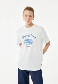 Witte katoenen t-shirt met blauwe grafische tekst "Sardine" en een illustratie van een sardineblik. Korte mouwen, ronde hals, relaxte fit.