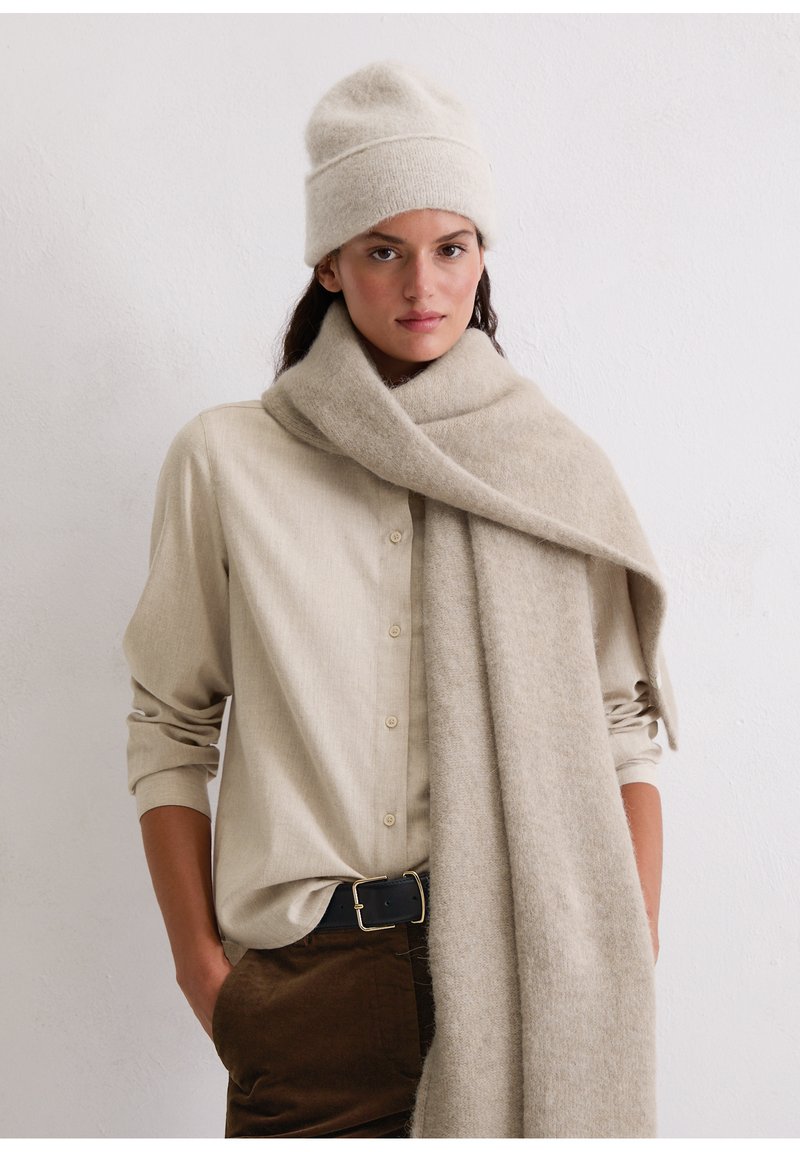 Beige wollen muts en lange sjaal gedrapeerd over een lichtbeige shirt. De sjaal heeft een zachte textuur en een eenvoudig, elegant ontwerp.