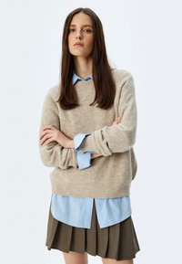 Maglione lavorato a maglia beige con scollo rotondo, indossato sopra una camicia blu chiaro con bottoni, abbinato a una gonna marrone plissettata.