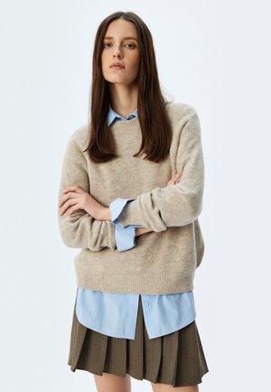 Beige gestrickter Pullover mit Rundhalsausschnitt, über einem hellblauen Hemd mit Knöpfen getragen, kombiniert mit einem plissierten braunen Rock.