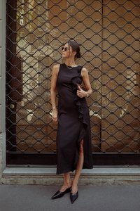 Musta olkaimeton mekko, jossa on sivupitkät röyhelöt, valmistettu teksturoidusta kankaasta. Yhdistetty mustiin kärkitossuihin ja pyöreisiin aurinkolaseihin verkkomaisen taustan edessä.